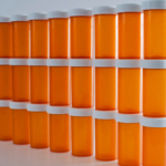 Three rows of identical orange prescription bottles with white lids and labels, arranged neatly in a line, increasing in height from left to right against a white background.