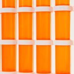 A close-up image of a collection of orange prescription bottles neatly aligned in rows, with white caps and blank labels, against a white background. The repetitive pattern and bright color highlight the pharmaceutical industry’s mass production of medication.